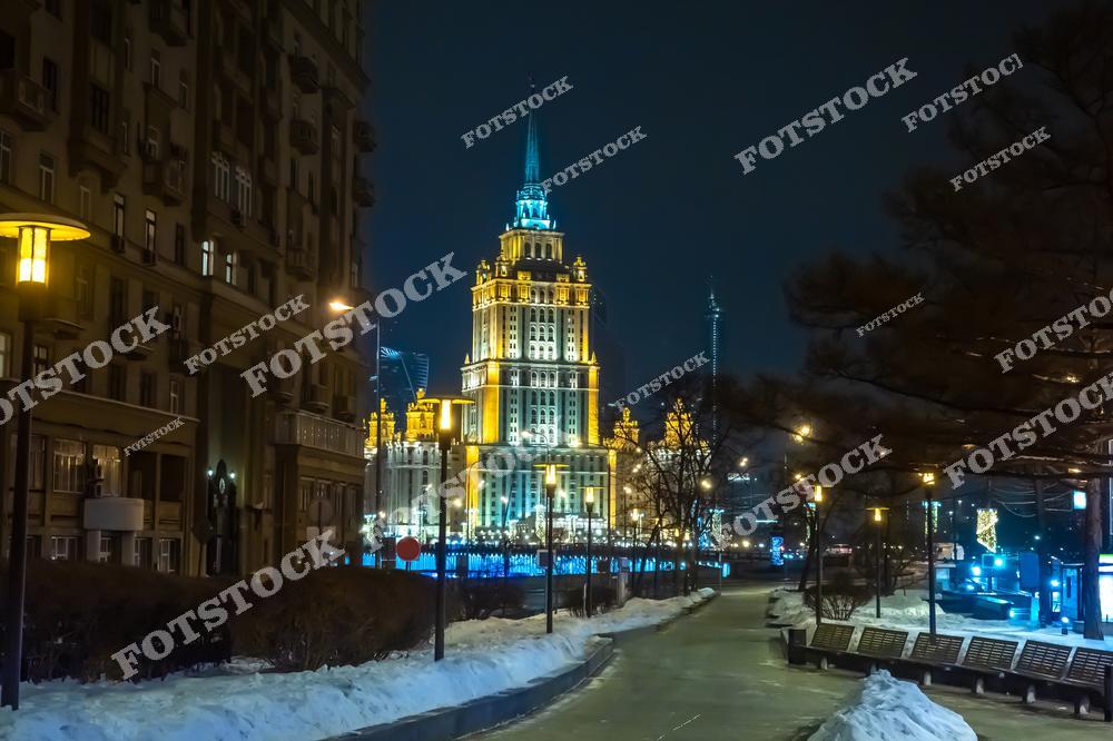 На фотографии изображена одна из сталинских высоток Москвы, освещённая огнями в ночное время. Здание контрастирует с окружающим пейзажем, который включает заснеженные деревья и улицы. Мягкий свет фонарей вдоль улицы и декоративные огоньки создают атмосферу спокойствия и зимнего уюта.