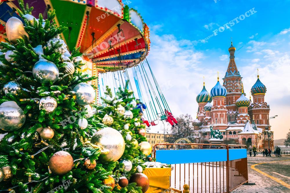 На фотографии запечатлена Красная площадь в Москве, украшенная к новогодним праздникам. Слева — яркие наряженные ёлки с золотыми и серебряными шарами, рядом с ними — красочная карусель с креслами на цепях. На заднем плане — величественный Собор Василия Блаженного с его знаменитой архитектурой и разноцветными куполами. Ясное голубое небо и солнечный свет делают зимний день особенно ярким. Вид передаёт атмосферу веселья, праздника и детского восторга. Пространство наполнено праздничным убранством и радостью зимнего сезона.
