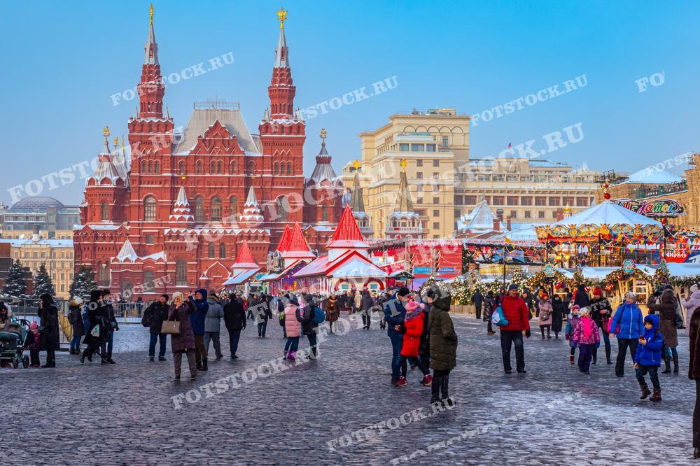 На фотографии изображена новогодняя ярмарка на Красной площади в Москве. Центральное место занимает ярмарка с палатками, украшенными к празднику. Вокруг карусели и павильонов прогуливаются люди, наслаждаясь зимним днем и праздничной атмосферой. На заднем плане виден Государственный исторический музей с его красными стенами и шпилями.