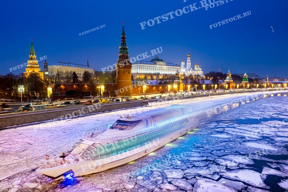 На фотографии изображен зимний вечер на Москве-реке с видом на Кремль. Прогулочный катер скользит по льду реки, создавая динамичный контраст с неподвижностью льдин. Кремлевские башни и здания красиво подсвечены, создавая волшебную атмосферу зимней Москвы.