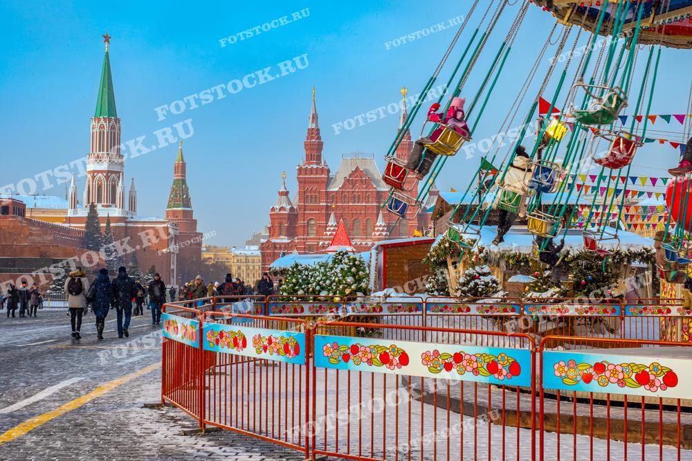 На фотографии изображена новогодняя карусель на Красной площади в Москве. На заднем плане виднеются Спасская башня Кремля и Государственный исторический музей, подчеркивая историческую значимость места. Люди прогуливаются по площади, наслаждаясь зимними развлечениями и праздничной атмосферой. Карусель украшена яркими цветами и гирляндами, создавая радостное настроение у посетителей.