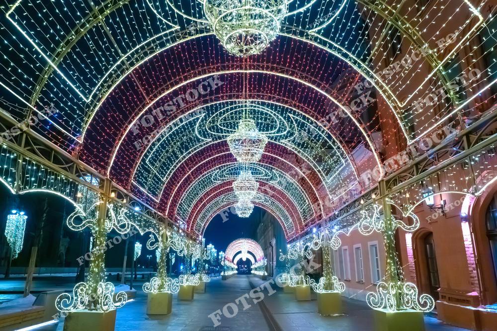 На фотографии изображен световой тоннель в Москве, украшенный праздничными гирляндами и световыми арками. Арки выполнены в золотисто-красной цветовой гамме и изящно подсвечены, создавая эффект сияющего коридора. На переднем плане видны детали тонкой светодиодной работы, а в перспективе тоннель уходит вдаль, придавая фотографии глубину.
