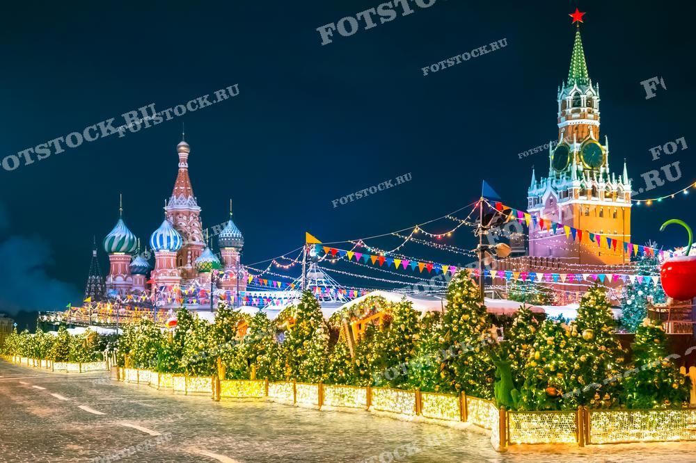 На фотографии изображена Красная площадь в Москве ночью, украшенная новогодней иллюминацией. В центре кадра видны Собор Василия Блаженного и Спасская башня, окруженные празднично украшенными ёлками и гирляндами. Яркие огни и декорации создают волшебную атмосферу праздника.