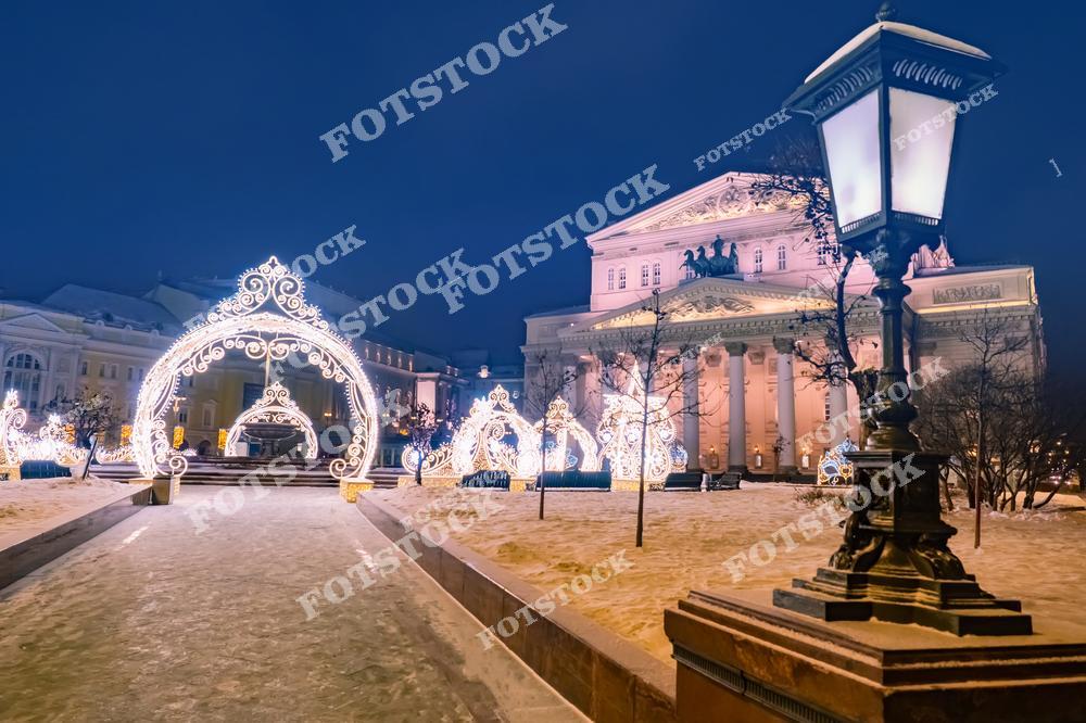 На фотографии изображён фасад Большого театра в Москве, освещённый теплым светом в зимний вечер. Перед зданием установлены световые арки и декоративные элементы, выполненные в новогоднем стиле. Площадь покрыта снежным слоем, а тропинки аккуратно расчищены. Слева и справа видны деревья с огнями и другие украшения, подчёркивающие атмосферу праздника. Лампа на переднем плане добавляет снимку уют и ощущение старинного городского ансамбля. Всё пространство наполнено светом, торжественностью и культурным духом Москвы.