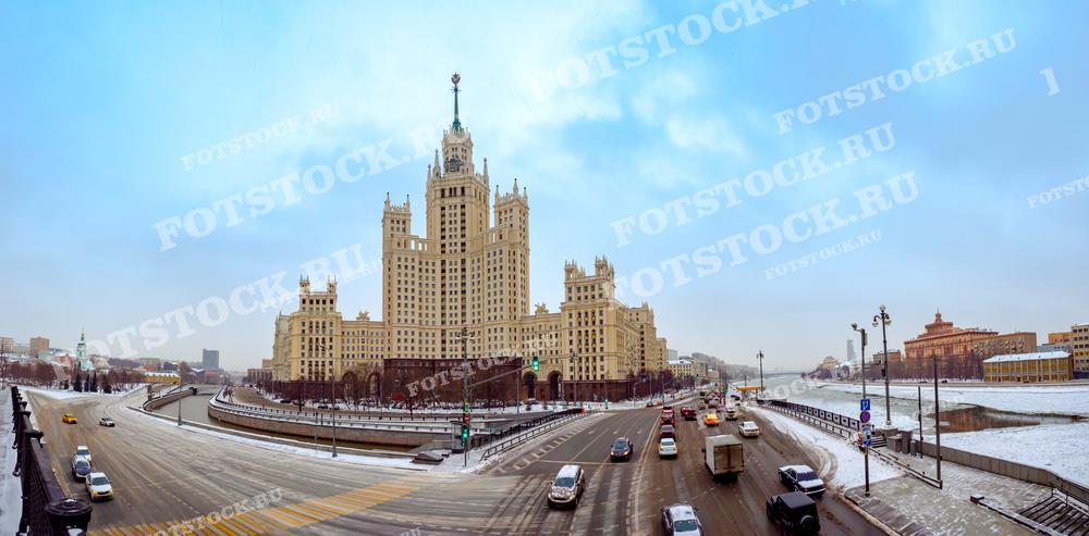 На фотографии изображена Котельническая набережная в Москве зимой. В центре кадра находится одна из сталинских высоток, величественное здание на фоне зимнего неба. По улицам движется транспорт, а вдоль набережной видна замерзшая Москва-река.
