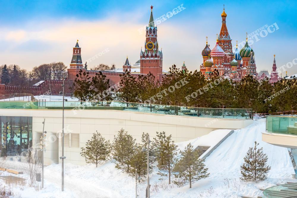 На фотографии изображен зимний вид на Кремль и Собор Василия Блаженного из парка Зарядье. На переднем плане можно увидеть современные архитектурные элементы парка, включая стеклянные конструкции и заснеженные холмы, окруженные хвойными деревьями. В центре кадра возвышается Спасская башня с курантами, а справа красочные купола Собора Василия Блаженного. Яркий зимний свет подчеркивает гармонию природы, современной архитектуры и исторического наследия в сердце столицы России.
