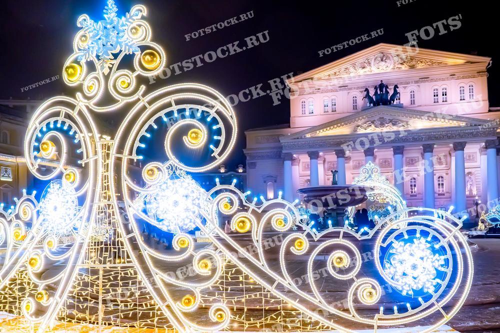 На фотографии изображен знаменитый Большой театр в Москве, одно из главных культурных достояний России. Перед театром установлены яркие новогодние световые инсталляции, которые добавляют площади особую праздничную атмосферу. Элегантные завитки из огней гармонично дополняют архитектуру театра, подчёркивая его классический стиль. Величественные колонны здания освещены мягким светом, а на фронтоне видна скульптура Аполлона на колеснице, создавая неповторимую композицию. Сцена выглядит особенно волшебно в преддверии новогодних праздников.