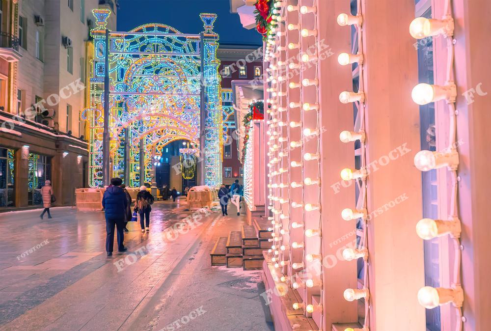 На фотографии изображена улица Москвы, украшенная к Новому году и Рождеству. По центру кадра возвышается большая световая арка, переливающаяся разноцветными огнями. Справа — фасад здания, украшенный гирляндами из лампочек в ретро-стиле. Люди гуляют по пешеходной зоне, наслаждаясь атмосферой праздника. Мокрая мостовая отражает огни и подчёркивает яркость оформления. Это одно из популярных мест среди горожан и туристов в зимние вечера.
