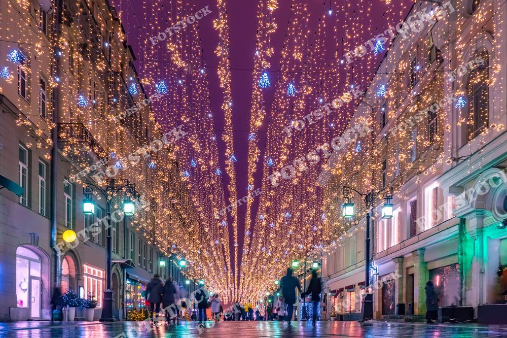 На фотографии изображена пешеходная улица Москвы, украшенная яркими новогодними гирляндами. Тысячи золотистых огней создают эффект звёздного неба, с вкраплениями голубых снежинок, добавляющих зимнее настроение. Люди неспешно гуляют, наслаждаясь праздничной атмосферой и теплом вечернего света. Фасады зданий, освещенные мягкими огнями, дополняют волшебный антураж улицы. Эта сцена передает настроение московских новогодних праздников и зимней красоты города.