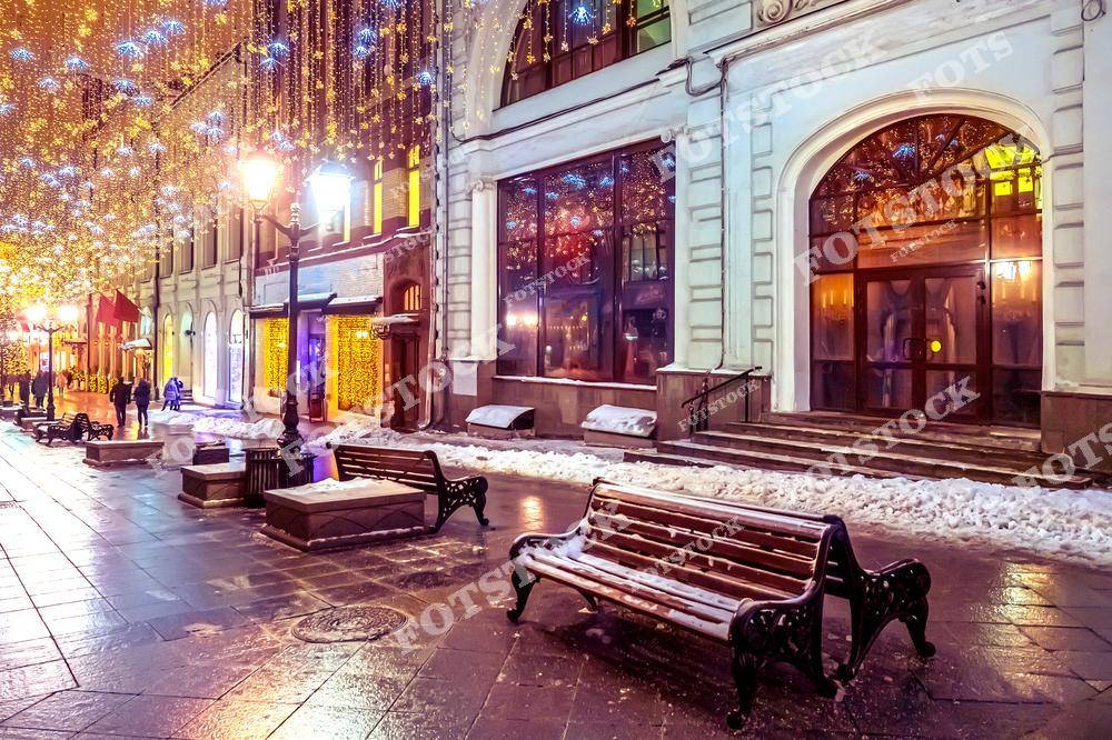 На фотографии изображена пешеходная улица Москвы, украшенная праздничными гирляндами. Над улицей сияют золотистые огоньки и световые снежинки, создавая сказочную атмосферу. Скамейки с декоративными элементами расположены вдоль фасадов исторических зданий, освещенных мягкими огнями. Легкий снег покрывает тротуары, добавляя зимнего уюта. Это место притягивает москвичей и туристов для вечерних прогулок в новогодний сезон, предлагая особую атмосферу спокойствия и праздника.