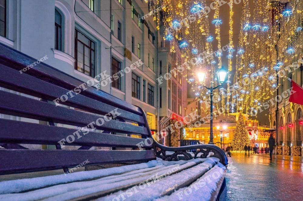 На фотографии запечатлена уютная московская улица в зимний вечер, украшенная праздничной иллюминацией. На переднем плане — покрытая снегом скамейка. Над улицей сияют золотые огоньки гирлянд с голубыми снежинками, освещая фасады зданий. На заднем плане видна большая праздничная елка, добавляющая торжественности композиции. Атмосфера улицы наполнена теплом, несмотря на зимний холод, и привлекает жителей и туристов для неспешных прогулок.