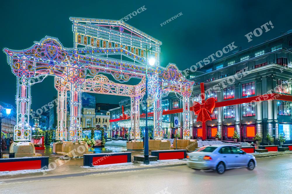 На фотографии запечатлена одна из московских улиц, украшенная к новогодним праздникам. В центре кадра находится яркая световая арка, выполненная в виде праздничной инсталляции с разноцветными огнями. На заднем плане видны здания с витринами, оформленными в виде подарков с красными лентами и бантами. На переднем плане — проезжающий автомобиль, добавляющий динамики к сцене. Зимняя ночь и снежные декорации создают атмосферу уюта и праздника в городской среде.
