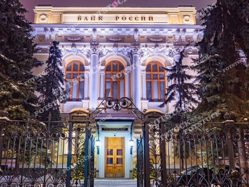 На фотографии изображено здание Центрального банка России, расположенное в Москве. Его фасад освещен мягкой подсветкой, подчеркивающей классические архитектурные детали, включая колонны, арочные окна и лепнину. Здание окружено хвойными деревьями, покрытыми легким слоем снега, что добавляет зимней сказочности. Элегантные кованые ворота на переднем плане завершают общий облик композиции. Это здание символизирует сочетание исторической архитектуры и современной городской среды.