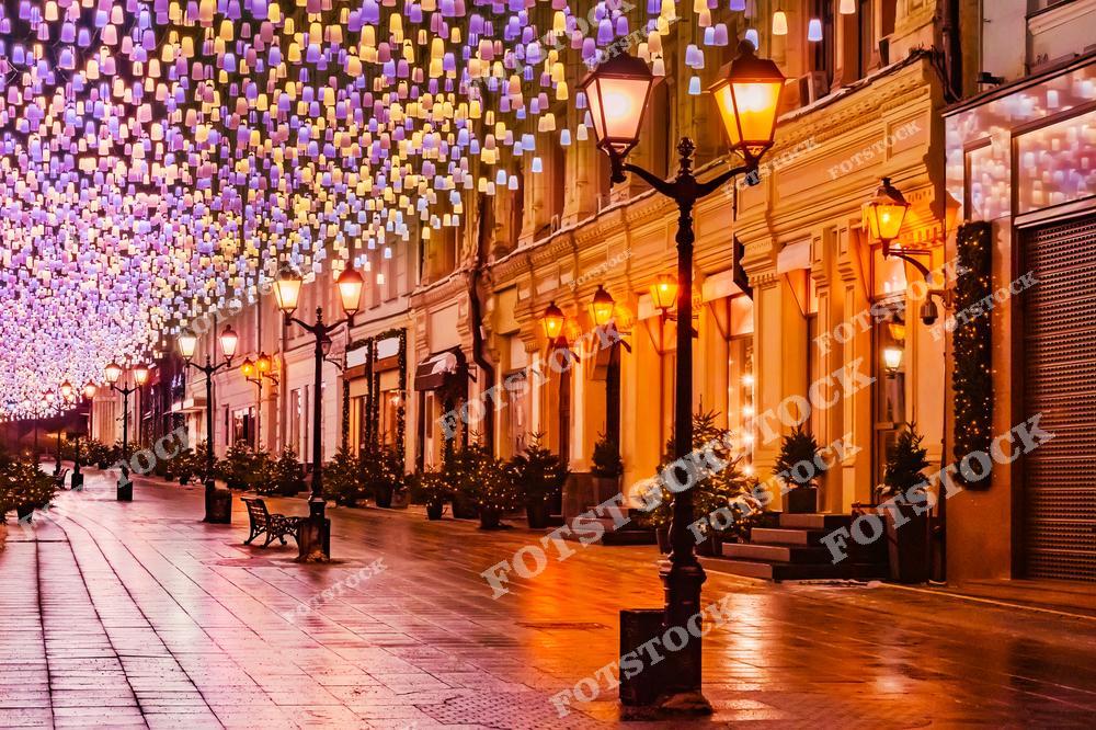 На фотографии изображена улица Москвы, украшенная яркими новогодними огнями. Улицу освещают фонари, а разноцветные огоньки создают атмосферу праздника и волшебства. Этот вид особенно красив в вечернее время, когда огни светятся на фоне городской архитектуры, создавая неповторимую атмосферу новогодних праздников.