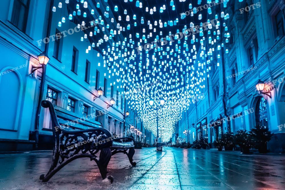 На фотографии изображена улица в Москве, украшенная множеством светящихся ламп, создающих ощущение звёздного неба над городом. Мягкое синее освещение зданий и отражение света на мокрой мостовой добавляют кадру волшебства и таинственности. Эта сцена является прекрасным примером городской иллюминации, делающей зимние вечера особенно уютными и праздничными.