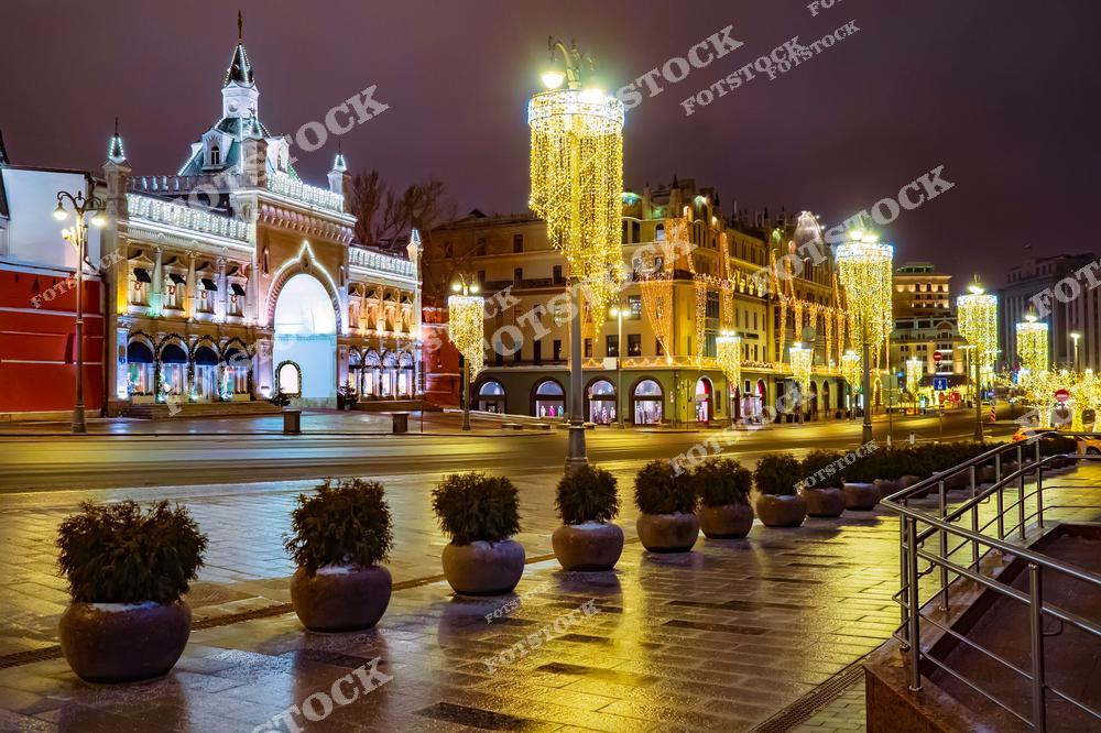На фотографии изображен празднично украшенный центр Москвы. Слева видно здание Исторического музея, украшенное белоснежной подсветкой и арками с гирляндами. Центральная улица озарена яркими световыми композициями в виде сверкающих цилиндров, подвешенных на фонарных столбах. Вдалеке можно рассмотреть другие исторические здания, гармонично вписывающиеся в праздничный антураж. Мокрая мостовая отражает огни, усиливая ощущение праздника и уюта зимнего вечера в столице.