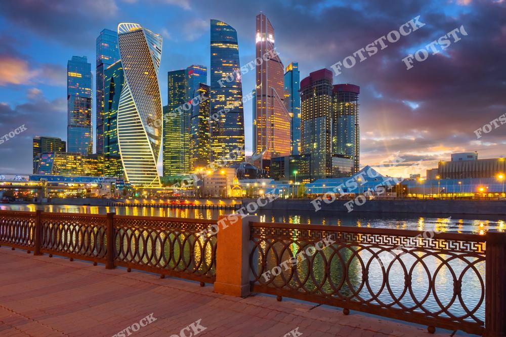 На фотографии изображён деловой центр Москвы — Москва-Сити, сияющий в лучах заката. Небоскрёбы, включая башни «Эволюция», «Федерация» и «Око», отражаются в воде Москва-реки. На переднем плане — ограждение набережной, выполненное в классическом стиле, создающее контраст с ультрасовременной архитектурой. Здание подсвечены разноцветными огнями, что усиливает эффект городской динамики. Небо окрашено в тёплые тона заходящего солнца, подчёркивая силуэт столицы. Это место стало символом нового облика мегаполиса.