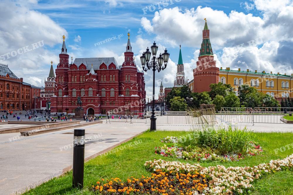 На фотографии изображен летний день у Красной площади в Москве. В центре кадра виден Государственный исторический музей и Кремлевские башни. На переднем плане находятся ухоженные цветочные клумбы и зеленый газон, создающие яркий контраст с историческими зданиями.