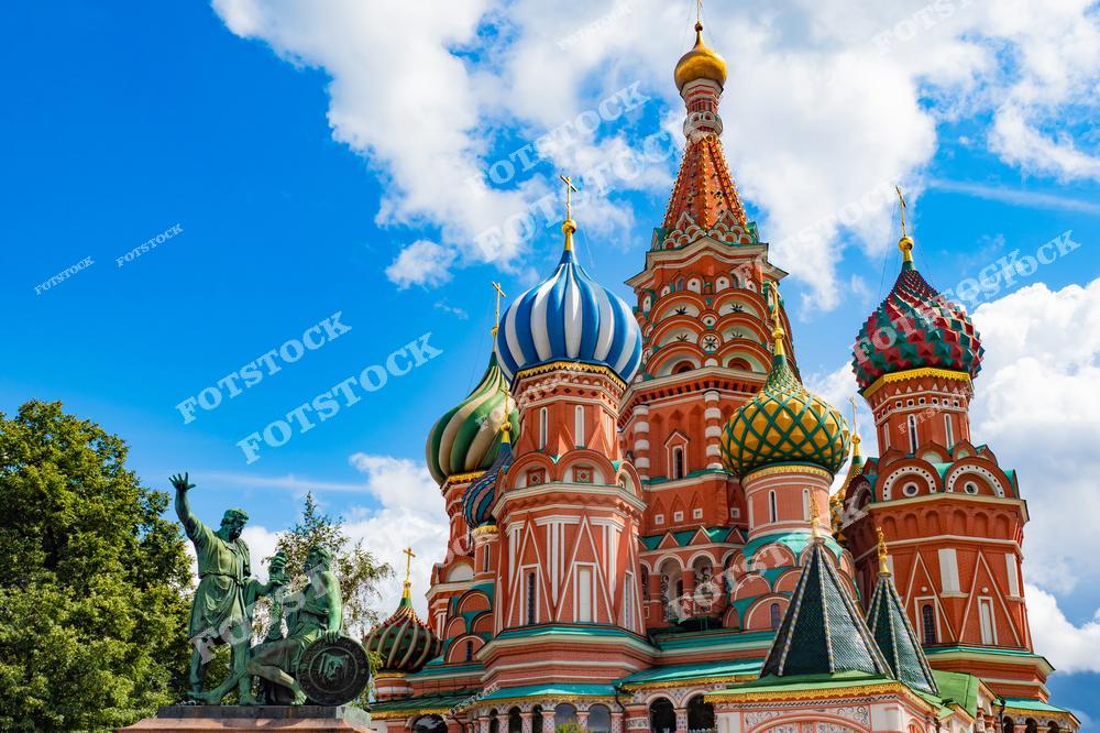 На фотографии изображен Собор Василия Блаженного с его яркими куполами, гармонично сочетающимися с голубым небом и белыми облаками. Слева расположен памятник Минину и Пожарскому, символизирующий героизм и единство народа. Купола собора, украшенные уникальными узорами, привлекают внимание своим разнообразием форм и цветов. Зеленые деревья на заднем плане добавляют свежести к летнему пейзажу. Эта сцена передает атмосферу исторической Москвы и её величия.