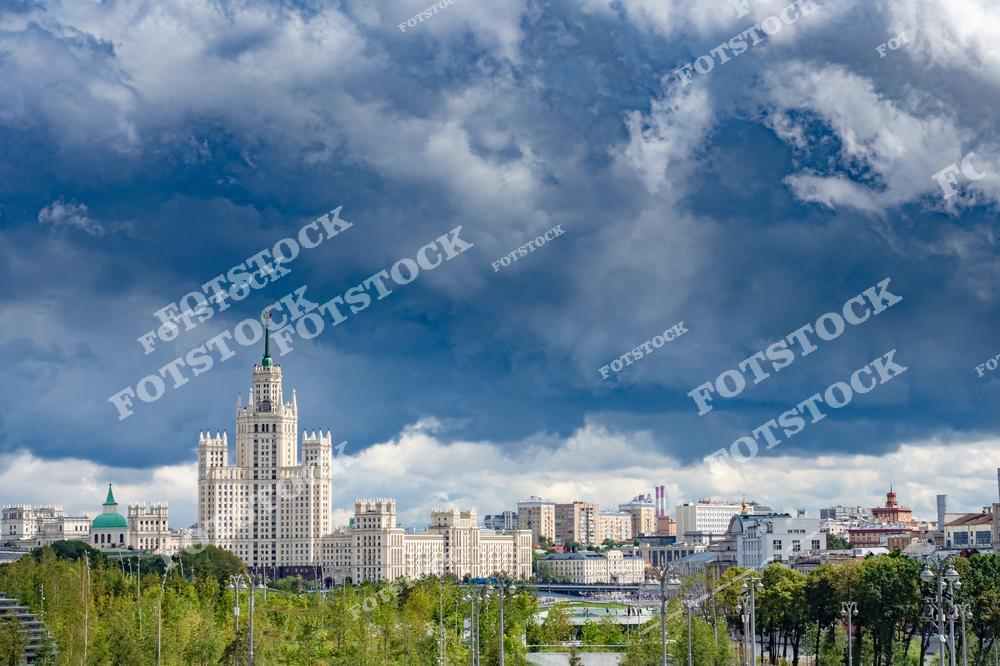 На фотографии изображена знаменитая сталинская высотка на Котельнической набережной. Её величественный фасад возвышается на фоне темных грозовых облаков, создавая впечатляющий контраст с ярко освещенными зданиями внизу. На переднем плане видна зелень парка Зарядье, добавляющая кадру природной свежести. Панорама включает современные и исторические элементы архитектуры Москвы, подчёркивая богатство её культурного и городского пейзажа. Эта сцена идеально отражает динамику столицы.