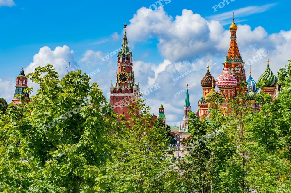 Красная площадь в Москве на солнечный летний день: зелёные деревья выделяются на переднем плане, за ними видны известные архитектурные достопримечательности — Спасская башня и храм Василия Блаженного с его яркими, цветными куполами. Небо голубое, с небольшими облаками.