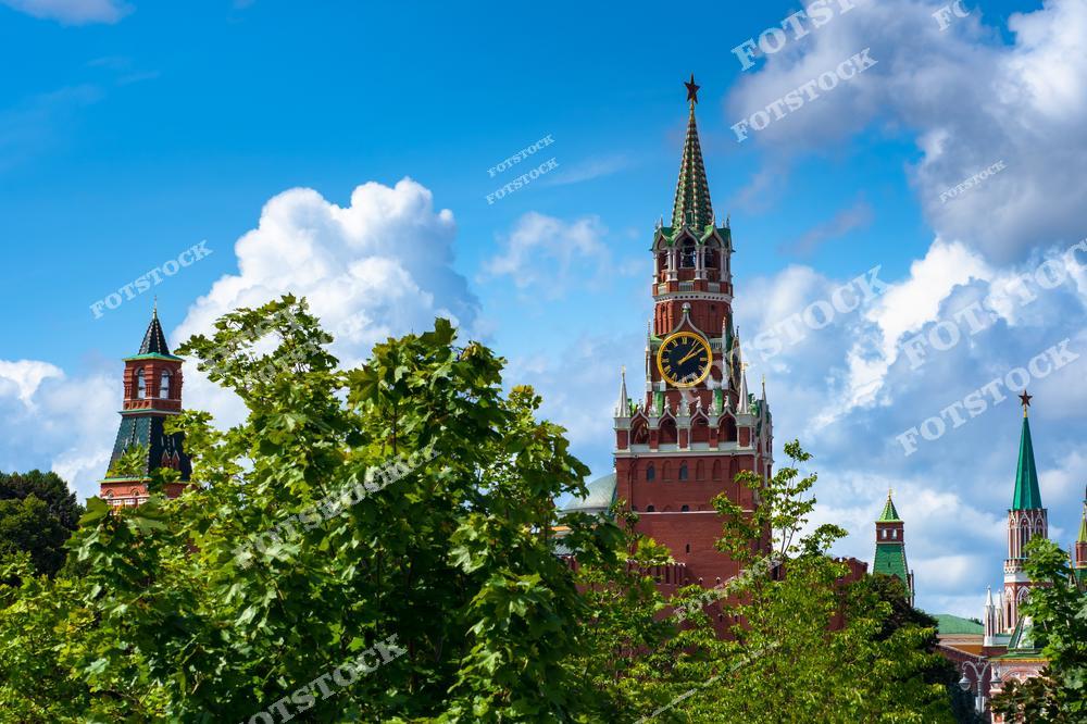 На фотографии изображена Спасская башня Московского Кремля — одна из самых узнаваемых достопримечательностей Москвы. Часы на башне показывают точное время, а золотая звезда сверкает на шпиле. На переднем плане — густая летняя листва деревьев, создающая ощущение природной свежести. Слева и справа видны другие кремлёвские башни с характерными зелёными крышами. Ясное голубое небо с облаками придаёт кадру яркость и лёгкость. Вид символизирует гармонию природы и исторической архитектуры в центре мегаполиса.