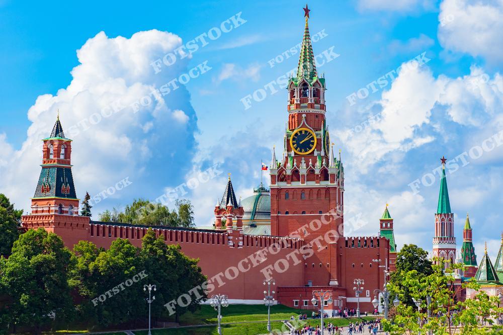 На фотографии изображена Спасская башня Московского Кремля с её знаменитым часами — курантами. Красные стены Кремля окружают башню, подчёркивая её величие и историческое значение. На заднем плане видны другие кремлёвские башни, создавая единый архитектурный ансамбль. Голубое небо с белыми облаками усиливает контраст между природой и архитектурой. Перед Кремлём можно увидеть людей, прогуливающихся по площади в летний день.