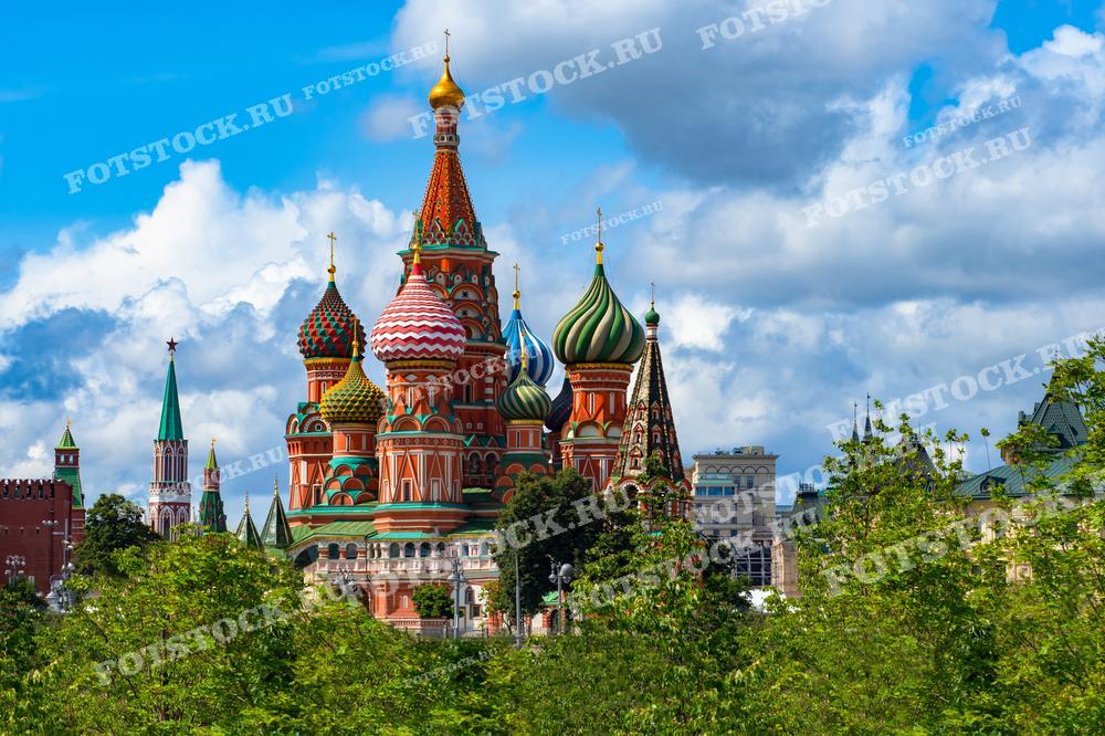 На фотографии изображен летний вид на Собор Василия Блаженного в Москве. Ветви деревьев на переднем плане обрамляют знаменитые купола собора и Кремлевские башни. Яркое голубое небо с облаками дополняет живописный городской пейзаж, подчеркивая красоту исторической архитектуры.
