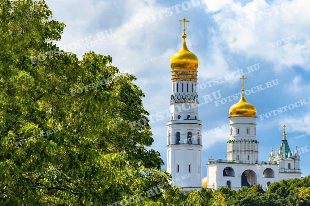 На фотографии изображена Колокольня Ивана Великого в Московском Кремле. Это дневной летний снимок, где золотые купола башен Кремля виднеются на фоне зелени деревьев и голубого неба. Фотография подходит для использования в туристических буклетах, статьях о Москве и иллюстрациях о достопримечательностях столицы.