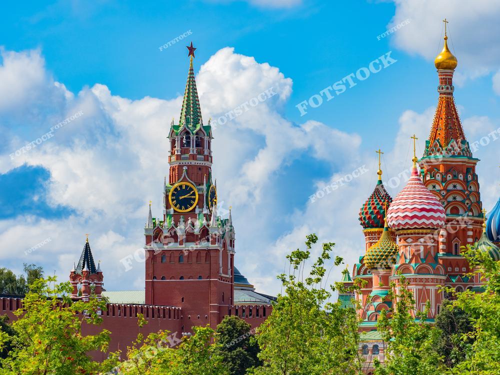 На фото запечатлен знаменитый московский Кремль, вид из парка Зарядье. На переднем плане — купола Собора Василия Блаженного, ярко выделяющиеся своей архитектурной красотой, а рядом — Спасская башня, одна из самых узнаваемых башен Кремля. Зелень деревьев парка Зарядье создает живописный контраст с величественными историческими зданиями. Этот вид является одной из самых популярных туристических достопримечательностей Москвы и символизирует сочетание истории и природы.