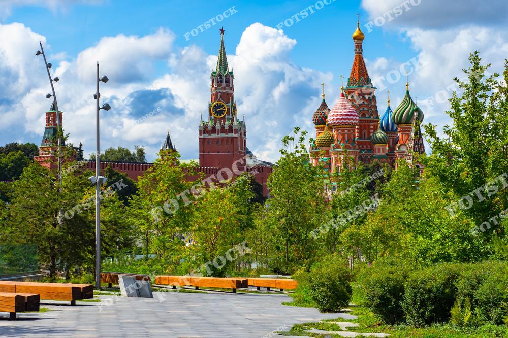 На фотографии изображены два главных символа Москвы — Собор Василия Блаженного с его яркими куполами и Спасская башня Кремля с часами. Эти архитектурные шедевры видны из парка Зарядье, где на переднем плане расположены зелёные насаждения и современные скамейки для отдыха. Голубое небо с белыми облаками подчёркивает яркость и контраст летнего дня. Сочетание природы и исторических памятников делает этот вид уникальным и привлекательным для москвичей и туристов.