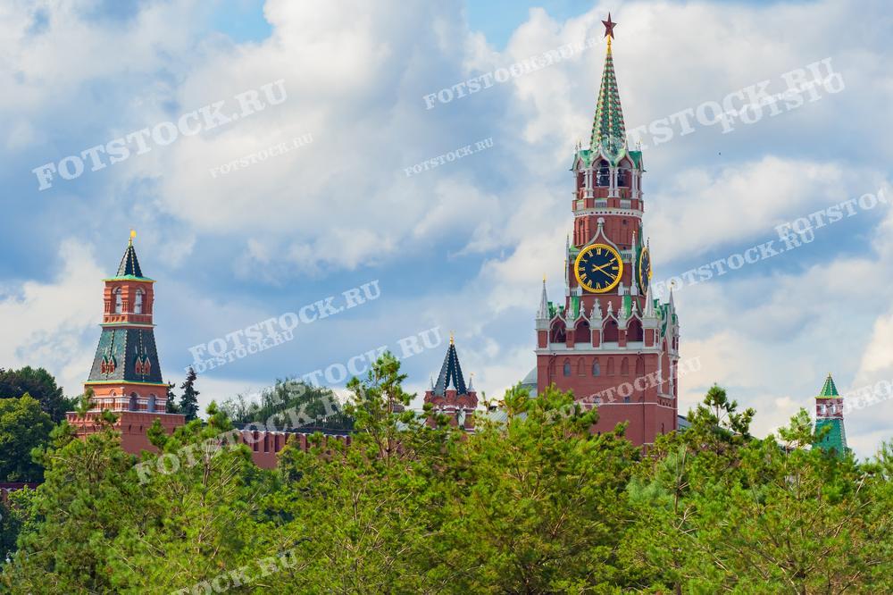 На фотографии изображен летний вид на Спасскую башню Кремля в Москве. Ветви деревьев на переднем плане создают зеленую рамку для башен Кремля. Яркое голубое небо с облаками подчеркивает величественность исторической архитектуры.