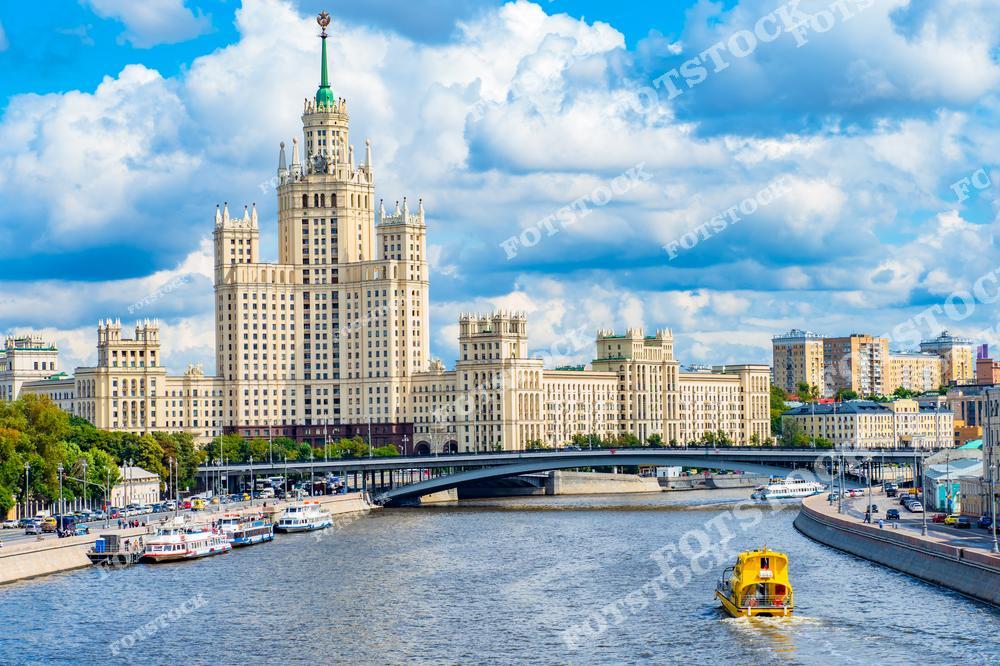 На фотографии изображена одна из знаменитых сталинских высоток, расположенная на Котельнической набережной. Здание возвышается на фоне голубого неба с белыми облаками, создавая величественный образ. На переднем плане проходит Москва-река, по которой плывут речные суда и ярко-жёлтая лодка, оживляющая кадр. Вид также включает мост, соединяющий берега, и архитектурные здания в окружении зелени. Это сочетание архитектурного наследия и динамичной городской среды идеально отражает жизнь Москвы.