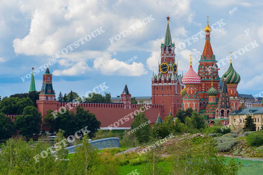 На фотографии изображен Кремль с его мощными красными стенами и башнями, включая Спасскую башню с часами. Рядом расположился Собор Василия Блаженного с его узнаваемыми разноцветными куполами. Вид открывается из парка Зарядье, где природа гармонично сочетается с историческими памятниками. Зелень деревьев и насыщенное голубое небо с облаками подчеркивают летний день. Этот кадр передает спокойствие и величие центра Москвы.