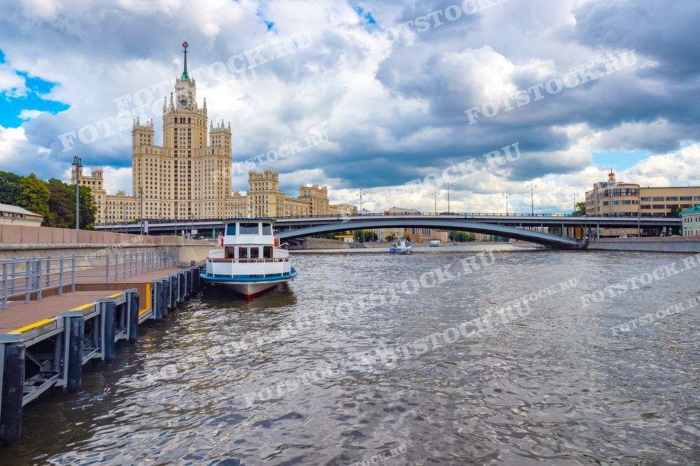 На фотографии изображена сталинская высотка на Котельнической набережной и Москва-река. Перед зданием виден мост и пришвартованный прогулочный катер.