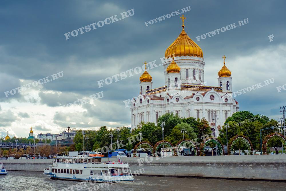На фотографии изображен Храм Христа Спасителя на фоне пасмурного или грозового неба. Впереди видна Москва-река с прогулочными катерами.