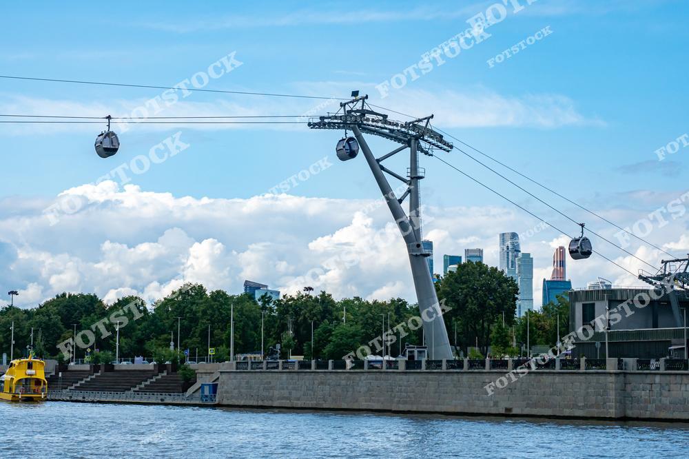 На фотографии изображена канатная дорога, проходящая над Москвой-рекой. Кабинки поднимаются над водой, предлагая великолепные виды на Москву. На заднем плане видны небоскрёбы Москва-Сити, подчеркивающие современный характер города.