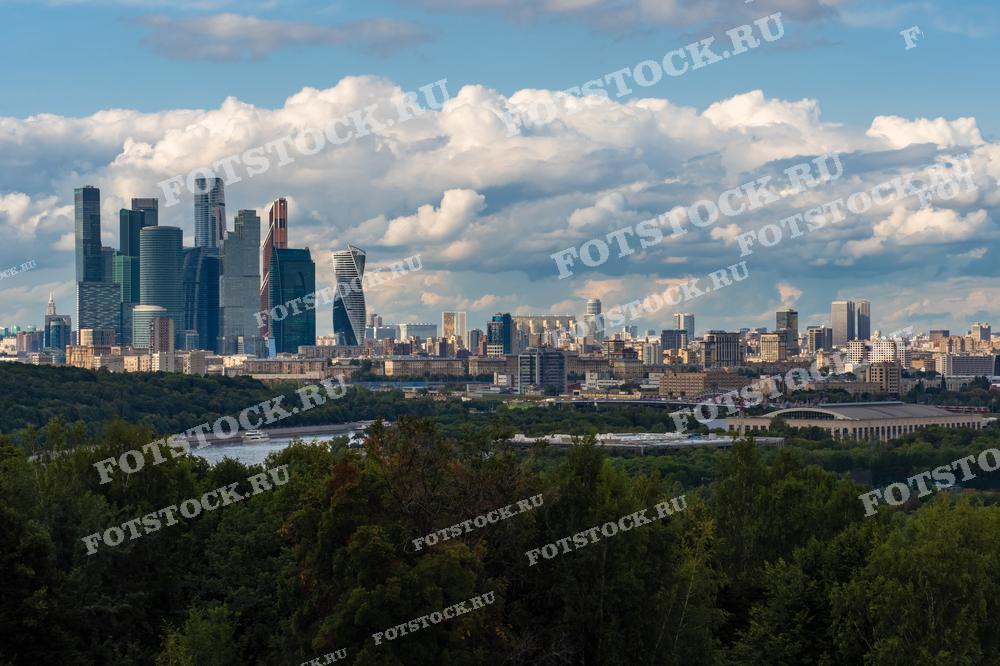 На фотографии изображен летний вид на Москва-сити и Москву-реку. Современные небоскребы делового центра возвышаются на фоне яркого летнего неба с облаками. Внизу видны зеленые зоны и парк, создающие контраст с городской застройкой.