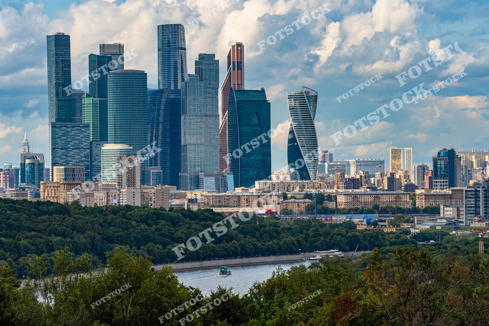 На фотографии запечатлен деловой центр Москвы — Москва-Сити. Высотные здания с зеркальными фасадами отражают голубое небо и облака, подчеркивая современность архитектуры. На переднем плане видна Москва-река, по которой плывет прогулочное судно. Окружающий городской пейзаж дополняет зелень парков и жилые дома.