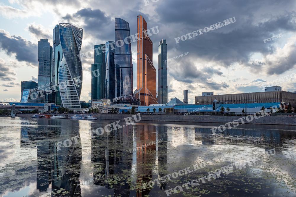 На фотографии изображена вечерняя панорама делового центра Москва-Сити и Москва-река. Небоскребы отражаются в спокойной воде реки, создавая впечатляющий вид на современную архитектуру Москвы.