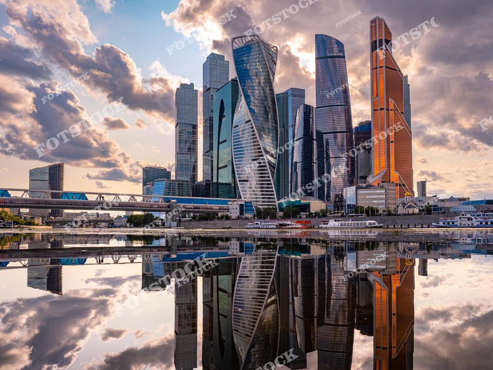 На фотографии представлены небоскрёбы делового центра Москва-Сити, отражающиеся в спокойной глади реки Москвы. Величественные здания, такие как башни "Федерация" и "Эволюция", подчеркивают современный облик столицы. На фоне закатного неба облака приобретают яркие и драматичные оттенки, добавляя картине романтичности. Пешеходный мост и причалы вдоль реки гармонично дополняют вид. Эта сцена демонстрирует симбиоз современной архитектуры и природной красоты.