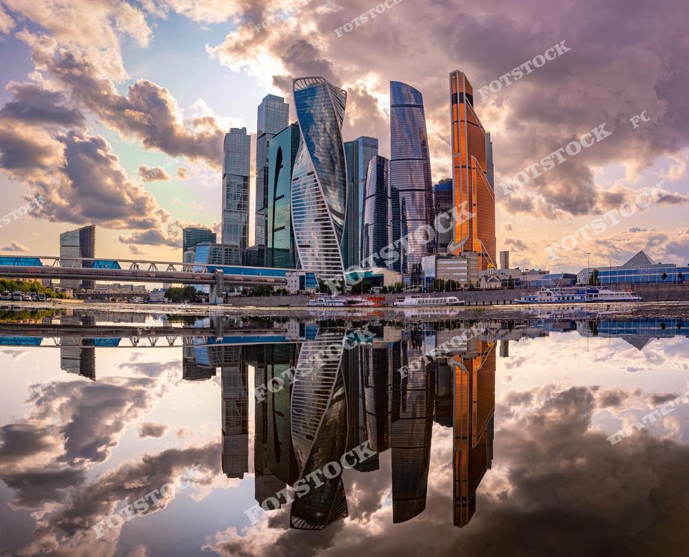 На фотографии изображены небоскрёбы делового центра Москва-Сити, высокие здания, отражающиеся в воде. Эти современные башни из стекла и бетона являются символом деловой активности столицы. Небоскрёбы, такие как "Восток", "Федерация" и другие, создают уникальный городской пейзаж, который привлекает как жителей, так и туристов. Строения на фоне облаков подчеркивают мощь и динамичность современной Москвы как центра бизнеса и экономики.
