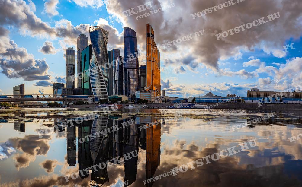 На фотографии изображен вид на деловой центр Москва-Сити, который отражается в водах Москвы-реки. Небоскрёбы с их уникальной архитектурой выделяются на фоне яркого неба с облаками. Отражение зданий в воде добавляет симметрии и глубины композиции, делая изображение идеальным для использования в фотоальбомах, рекламных материалах, а также в статьях о современной архитектуре и градостроительстве.