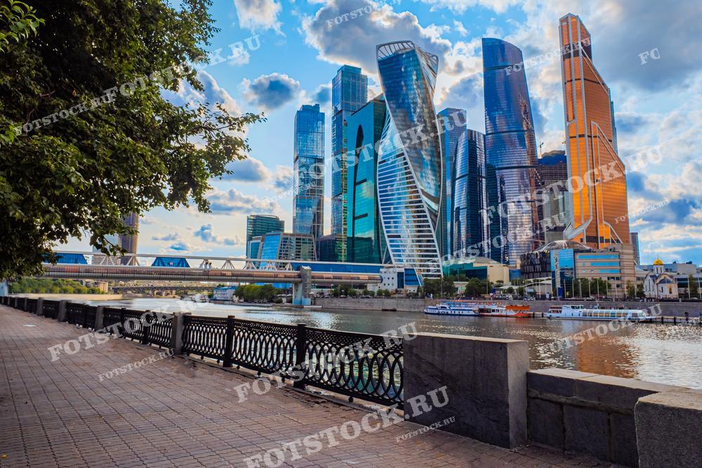 На фотографии изображены небоскрёбы Москва-Сити, расположенные на берегу реки Москва. Современные здания отражают свет летнего дня, а их стеклянные фасады блестят на солнце. На переднем плане видна набережная с прогулочными дорожками, где можно насладиться видом на реку и город. Река Москва оживлена прогулочными судами, которые подчёркивают активный ритм столицы. Высотные здания Москва-Сити символизируют финансовую и деловую мощь города.
