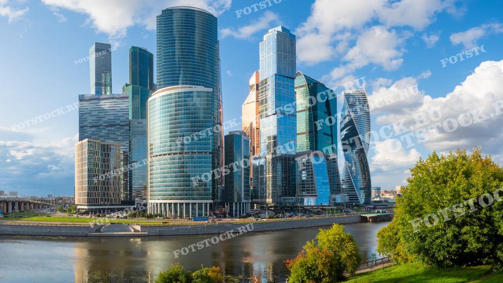 На фотографии изображен дневной вид на деловой центр Москва-Сити и Москву-реку. Современные небоскребы отражаются в воде реки на фоне ясного неба, создавая впечатляющий вид на Москву.