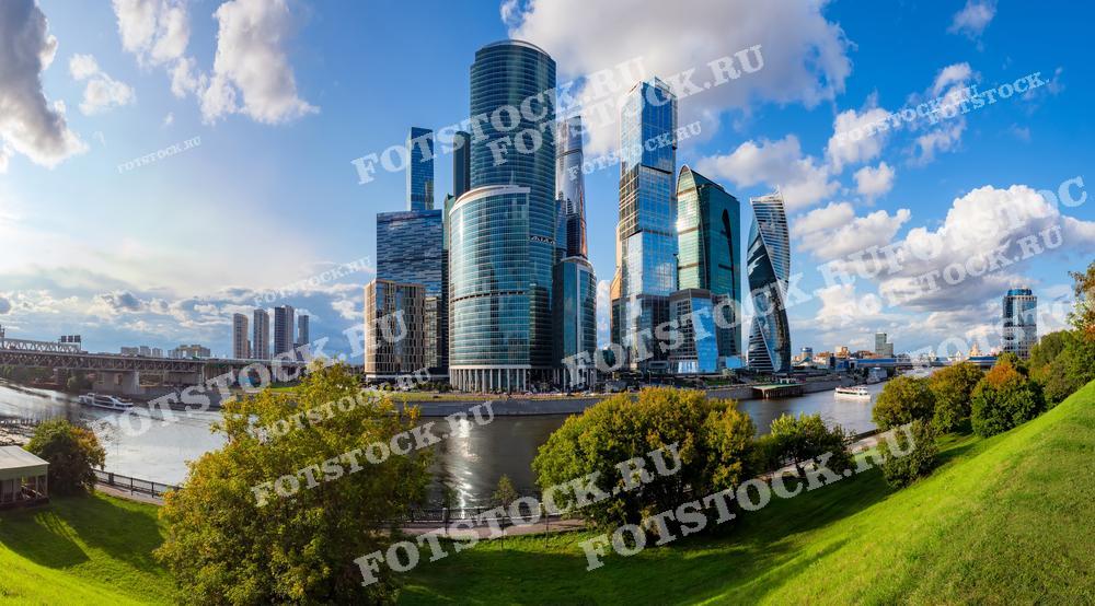 На фотографии изображен деловой центр Москва-Сити, состоящий из современных небоскребов, отражающих свет голубого неба. В кадре также видна Москва-река, вдоль которой тянутся зелёные парки и набережные. Офисные здания из стекла и стали символизируют динамичное развитие столицы. Справа от центра снимка выделяются башни "Эволюция" и "Федерация", одни из самых высоких зданий в России.