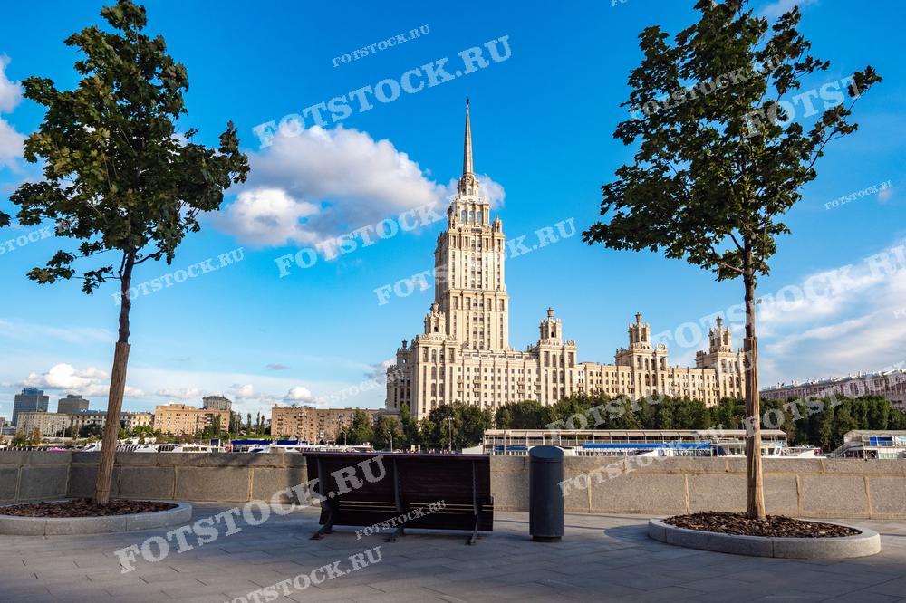 На фотографии изображена одна из знаменитых сталинских высоток в Москве. Здание выделяется своей величественной архитектурой и является одним из символов столицы. Фотография сделана с современной обзорной площадки, где чистые линии городского дизайна в сочетании с зеленью создают контраст с историческим зданием. Этот снимок идеально подходит для использования в культурных, архитектурных и туристических публикациях, а также может служить примером сочетания исторической архитектуры и современного городского развития.