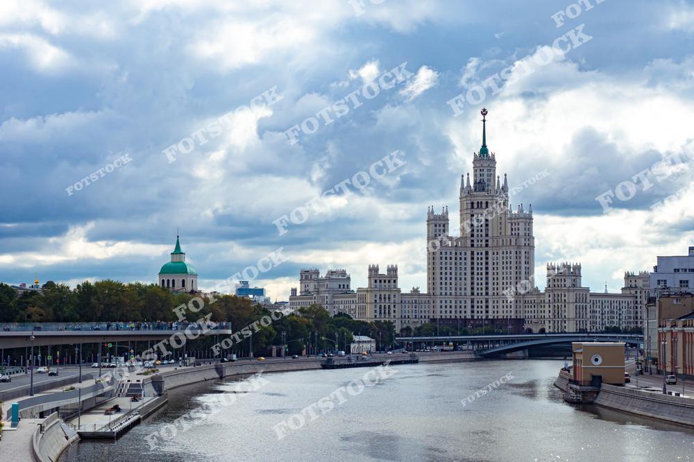 На фотографии изображен вид на Москву в пасмурный день с Москвы-реки. На переднем плане — высотное здание на Котельнической набережной, одно из известных сталинских небоскрёбов, которое является частью исторической архитектуры города. Вдали виден Парк Зарядье с пешеходной платформой, которая предлагает уникальный вид на столицу. Этот пейзаж является визитной карточкой Москвы, демонстрируя сочетание истории, природы и современной урбанистики.