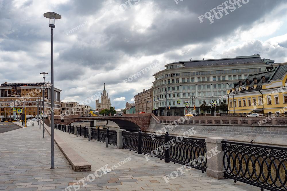 На переднем плане изображён мост через канал в центре Москвы, украшенный изящными коваными перилами. На заднем плане виднеется сталинская высотка на Котельнической набережной, одна из семи знаменитых высоток Москвы. Современные и исторические здания формируют гармоничный городской пейзаж. Мощёные тротуары и уличное освещение подчёркивают благоустройство района. Облачное небо добавляет фотографии глубину и драматизм, подчеркивая уникальность столичной архитектуры.