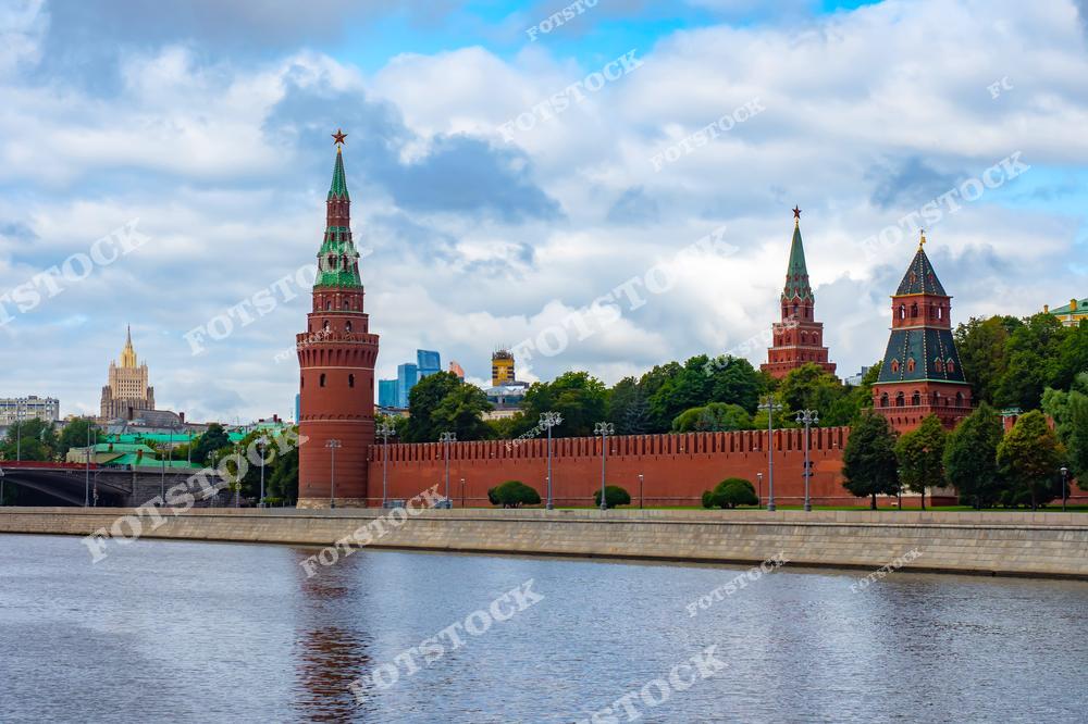 Изображение Московского кремля, вид с реки Москвы в ясный день. Красные стены и башни Кремля, увенчанные зелёными куполами, доминируют в композиции. Вдалеке видны городские здания, покрытие небольшими облаками на фоне голубого неба. На переднем плане тихая гладь реки отражает архитектурные детали.